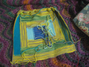 A knitted square in a quilt pattern known as log cabin, this one made with sock yarns and consisting of bright yellow, green, teal, aqua, and blue/purple strips. This square has stitches picked up around three sides, which will form the sides and bottom of a bag.