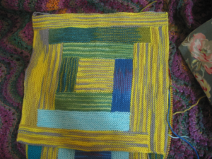 A knitted square in a quilt pattern known as log cabin, this one made with sock yarns and consisting of bright yellow, green, teal, aqua, and blue/purple strips.